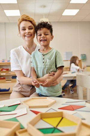 lächelnder Lehrer umarmt afrikanisches amerikanisches Kind in der Nähe von Unterrichtsmaterialien in der Montessori-Schule