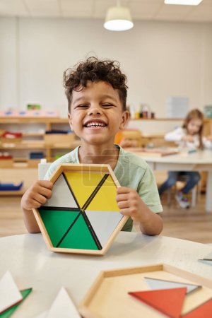 Foto de Feliz afroamericano chico celebración de juego de madera cerca de mesa en clase en montessori escuela - Imagen libre de derechos