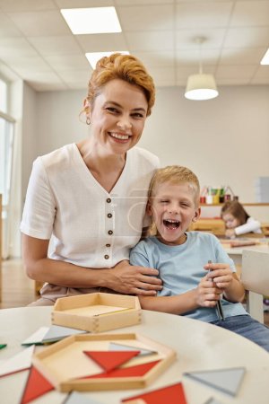 fröhlicher Lehrer umarmt Jungen in der Nähe von Holzspiel auf dem Tisch in der Montessori-Schule im Hintergrund
