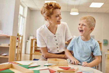 carefree teacher hugging boy near wooden game on table in montessori class