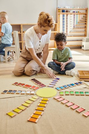 Photo for Teacher talking to african american boy near colorful wooden bricks on floor in montessori school - Royalty Free Image