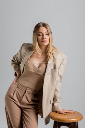 pretty young blonde woman in formal beige attire leaning on chair on grey backdrop, fashion concept