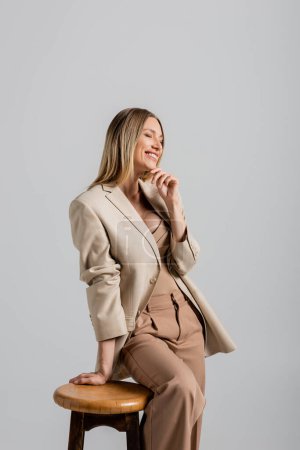 cheerful blonde woman in formal suit leaning on chair and touching chin on grey backdrop, fashion