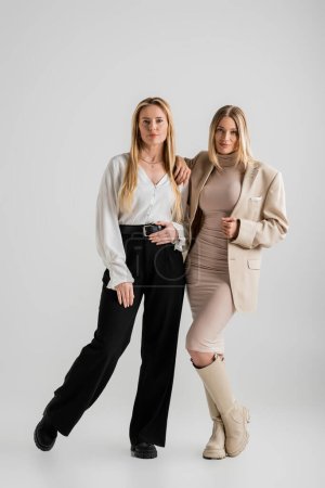 elegantes hermanas rubias en traje formal posando juntas sobre fondo gris, concepto de moda
