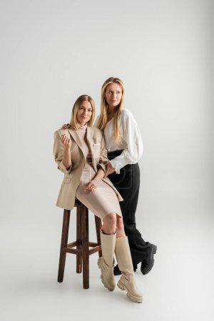two elegant stylish sisters posing on grey backdrop one sister sitting on chair, bonding, fashion