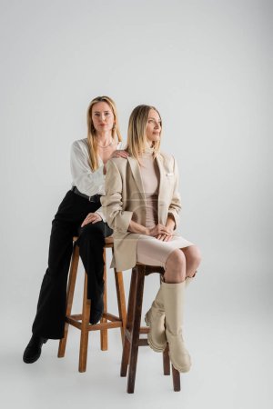 attractive stylish sisters in formal wear sitting on chairs, hand on shoulder, fashion and style