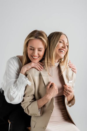 Photo for Beautiful stylish sisters in formal outfits beaming sitting on chairs and hugging, fashion concept - Royalty Free Image