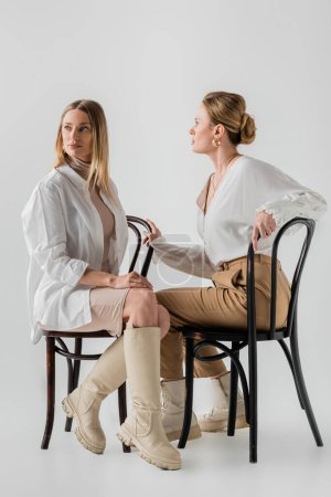 two attractive blonde sisters in attire sitting on chairs and looking away, bonding, fashion