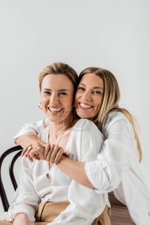 portrait of elegant blonde sisters sitting on chairs hugging and smiling, style and fashion