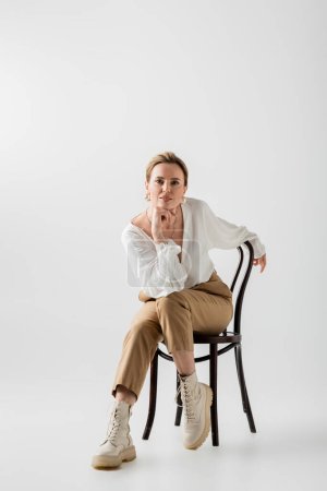 voguish hübsche Frau sitzt auf Stuhl und blickt in die Kamera, Hand zum Kinn, Stil und Mode