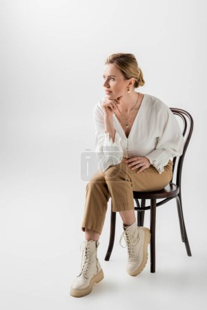 Blonde Frau in pastellfarbener Kleidung sitzt auf Stuhl und schaut weg, Hand an Kinn, Stil und Mode