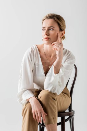 schöne blonde Frau in formeller Kleidung sitzt auf einem Stuhl und berührt ihr Gesicht, Stil und Mode