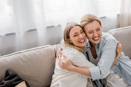 glückliche Schwestern in lässigen Outfits, die auf dem Sofa sitzen und umarmen und lächeln, Qualitätszeit, familiäre Bindung