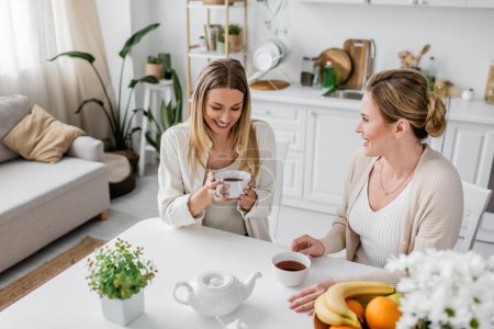 dos atractivas hermanas felices con atuendo casual sentadas en la mesa bebiendo té y sonriendo, uniéndose