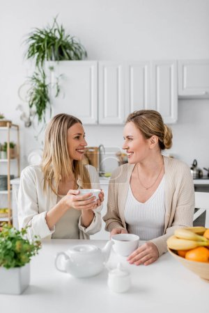 Photo for Close up two sisters sitting at table drinking tea and smiling to each other, family bonding - Royalty Free Image