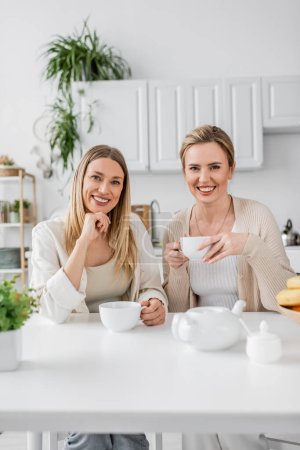 zwei Schwestern in pastellfarbenem Outfit, die lächelnd Tee trinken und in die Kamera schauen, Familienbande