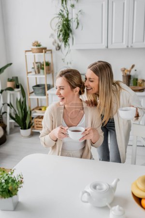 close up cheerful sisters in casual attire smiling and looking away with kitchen backdrop, bonding