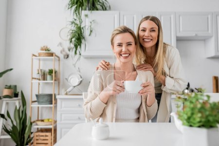 zwei blonde Schwestern trinken Tee und lächeln auf Küchenhintergrund mit Pflanzen, familiäre Bindung