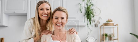 Photo for Blonde sisters smiling and looking at camera on kitchen backdrop with plants, family bonding, banner - Royalty Free Image