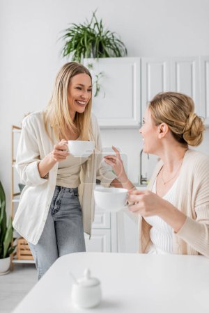 Photo for Two attractive sisters smiling and looking at each other on kitchen backdrop with plants, bonding - Royalty Free Image