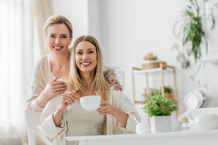 two cheerful pretty sisters looking at camera smiling and posing on kitchen backdrop, family bonding