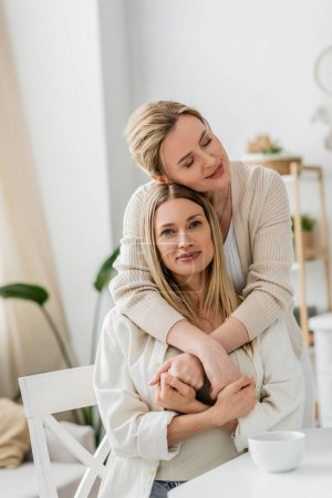 zwei blonde trendige Schwestern lächelnd und umarmend auf Küchenkulisse mit Pflanzen, familiäre Bindung