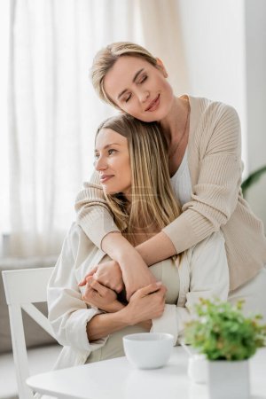 portrait of attractive blonde sisters hugging and holding hands on kitchen backdrop, family bonding