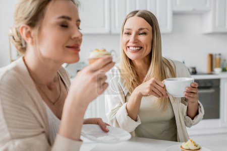 zwei schöne Schwestern in lässiger Kleidung, die Cupcakes probieren und Tee trinken, Familienbande
