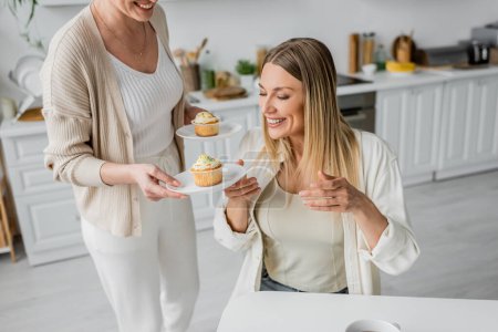 cropped view of two sisters looking at cupcakes and smiling on kitchen backdrop, family bonding