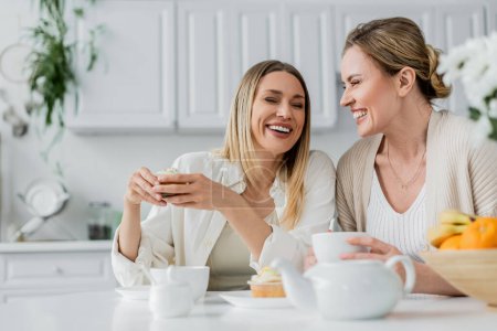 glückliche blonde Schwestern lachen am Tisch und genießen Tee und Cupcakes, Zweisamkeit, familiäre Bindung