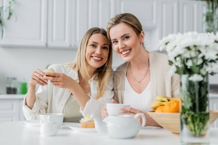 Photo for Cheerful lovely sisters in trendy attire enjoying tea and cupcakes and looking at camera, bonding - Royalty Free Image