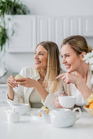 two good looking blonde sisters enjoying cupcakes and tea and looking away, pointing finger, bonding