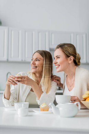 Téléchargez les photos : Chics soeurs blondes dans cardigans pastel savourant cupcakes et thé et détournant les yeux, liens de famille - en image libre de droit