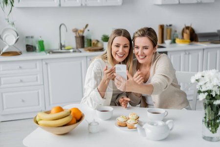 zwei Schwestern in pastellfarbener Kleidung machen Selfie am Küchentisch mit Cupcakes und Tee, Familienbande