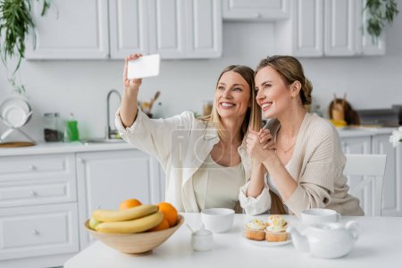 Téléchargez les photos : Sœurs chics dans les cardigans pastel à la mode faire du selfie à table dans la cuisine, liens familiaux - en image libre de droit