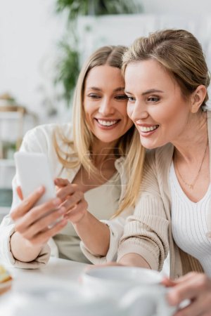 Téléchargez les photos : Fermer deux sœurs dans de belles tenues regardant le téléphone mobile et souriant, liens de famille - en image libre de droit