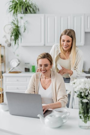 zwei blonde Schwestern in trendiger Kleidung schauen auf Laptop auf Küchenhintergrund, familiäre Bindung