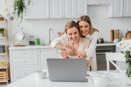 Foto de Atractivas hermanas rubias mirando a la computadora portátil sonriendo y abrazándose por detrás, vinculación familiar - Imagen libre de derechos