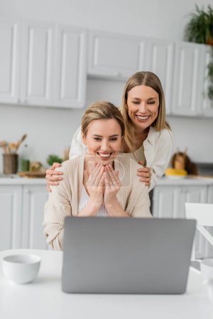 Lächelnde Schwestern in schönen lässigen Strickjacken mit Blick auf Laptop, hochwertige Zeit und familiäre Bindung