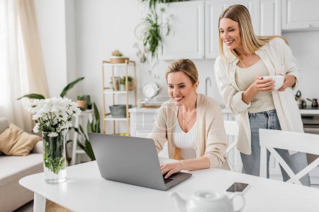 Photo for Good looking cheerful sisters in lovely attire looking at laptop and holding a tea cup, bonding - Royalty Free Image