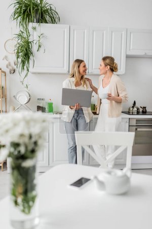 atractivas hermanas rubias de pie en la cocina con el ordenador portátil mirándose, vinculación familiar