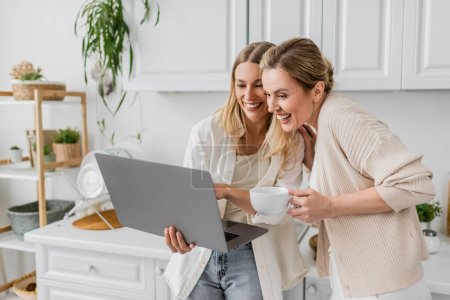 zwei fröhliche Schwestern, die auf Laptop schauen und aufrichtig lächeln, Teetasse in der Hand, Familienbande