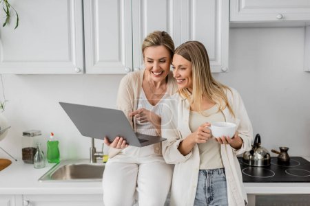 Photo for Attractive jolly sisters in lovely outfit looking at laptop on kitchen furniture backdrop, bonding - Royalty Free Image