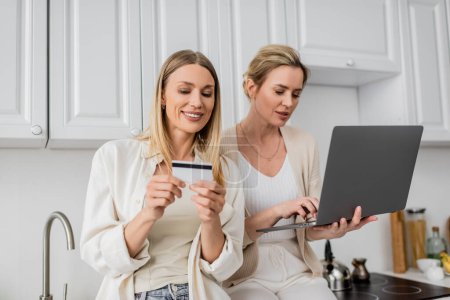 hermanas bonitas y guapas en atuendo pastel de moda sosteniendo tarjeta de crédito y computadora portátil, vinculación familiar