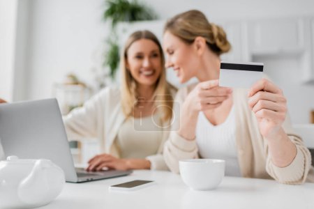 two smiling attractive sisters at kitchen table looking at laptop and holding credit card, bonding