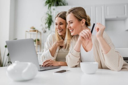 two cheerful blonde sisters holding credit card looking at laptop and smiling sincerely, bonding
