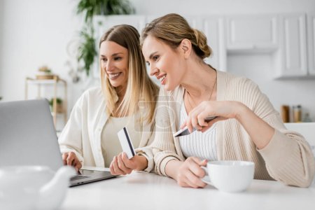 zwei hübsche blonde Schwestern in trendiger Kleidung mit Kreditkarte und Teetasse, die auf Laptop schauen,
