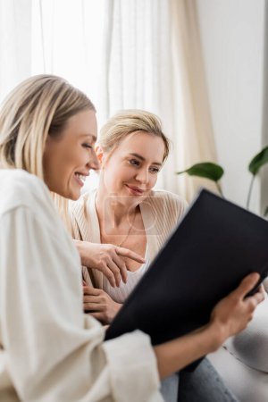 Photo for Two smiley attractive sisters in trendy casual cardigans looking at photo book, family bonding - Royalty Free Image