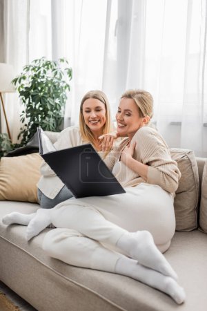 two smiling blonde sisters looking at photo album on white curtains backdrop, family bonding