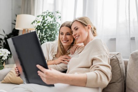 Photo for Pretty young sisters hugging and looking at photo book while sitting on sofa, family bonding - Royalty Free Image